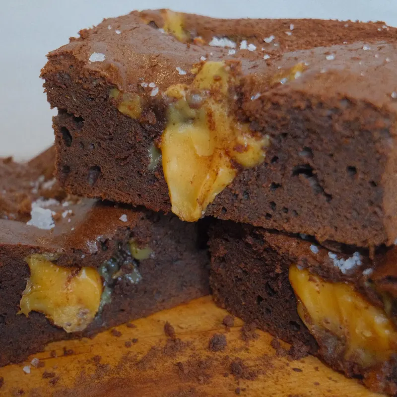 Stacked fudgy brownies with pockets of salted caramel and flakes of sea salt on a wooden board