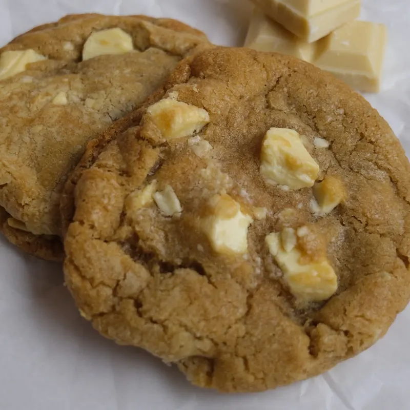 Two thick white chocolate chunk cookies on baking paper beside a few squares of white chocolate